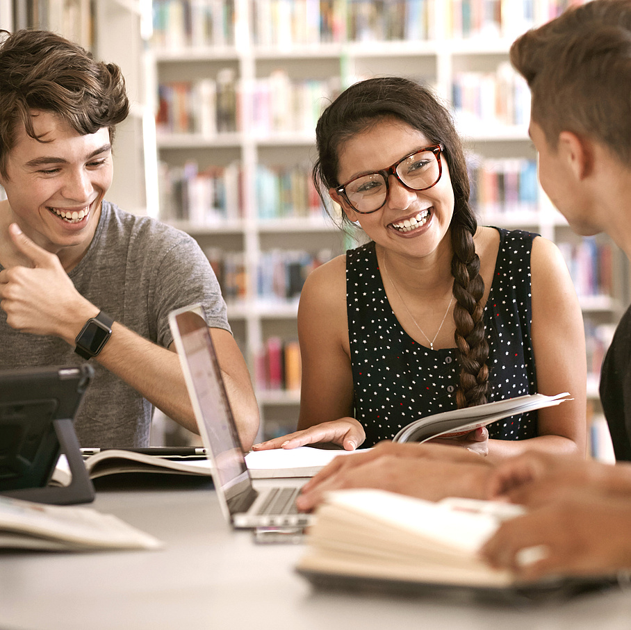 3 students at a table in front of a bookshelf.