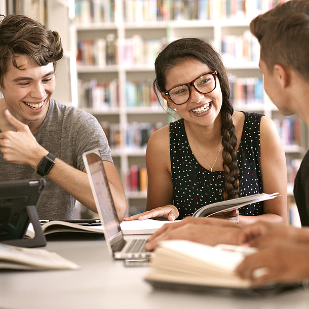 3 junge Leute sitzen an einem Tisch vor einem Bücherregal.