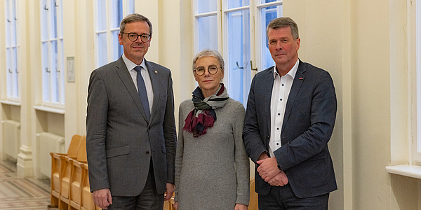 Drei Personen blicken in die Kamera, links und rechts stehen zwei Männer, in der Mitte eine Frau. Sie sind formal gekleidet.