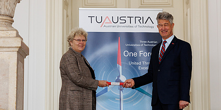 Eine Frau reicht einem Mann ein Staffelholz als Zeichen der Staffelübergabe der TU Austria Präsidentschaft