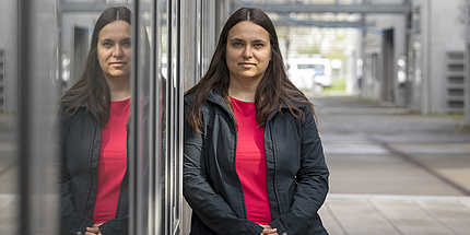 A brown haired women wearing a red shirt and black jacket is leaning against a wall.