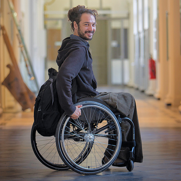 A man sits in a hallway of the TU Graz in a wheelchair. He is smiling at the camera.