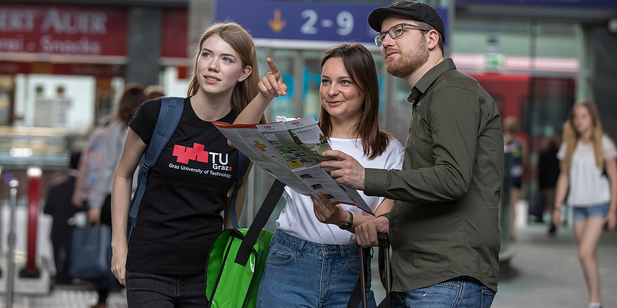 Two female students one of whom is pointing and one male student stand at the railway station and orientate themselves using a map.