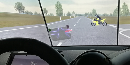 An intersection on an open country road is shown from the cockpit perspective; a yellow motorcycle is approached from the right; a display is to be found on the windscreen showing the intersection and the motorcycle as a red dot; the intersection is also shown virtually on the instrument panel behind the steering wheel. 