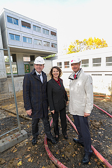 2 Männer und eine Frau stehen auf einer Baustelle. Die beiden Männer tragen einen weißen Helm. Alle drei blicken in die Kamera. 