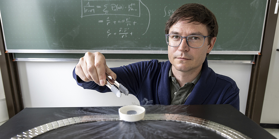 A man sits in front of a blackboard and looks into the camera. He holds a pair of pliers over a small white container.