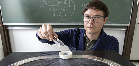 Ein Mann sitzt vor einer Tafel und blickt in die Kamera. Er hält eine Zange über einem kleinen, weißen Gefäß.