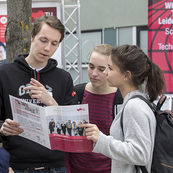 Ein junger Mann zeigt zwei Schülerinnen den Weg am Campus der TU Graz.