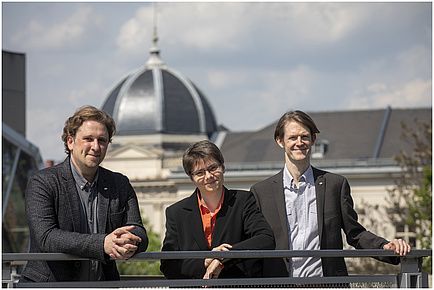 Two men, left with grey shirt, right with blue shirt, in the middle a woman with orange blouse stand in on a roof terrace in the background you can see a dome.