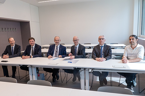Sechs Männer sitzen an einem Tisch und blicken in die Kamera.