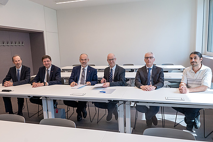 Sechs Männer sitzen an einem Tisch und blicken in die Kamera.