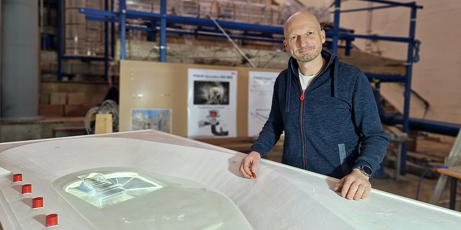 [Translate to Englisch:] Ein Mann mit blauer Weste steht neben einem Plexiglasmodell und schaut in die Kamera.