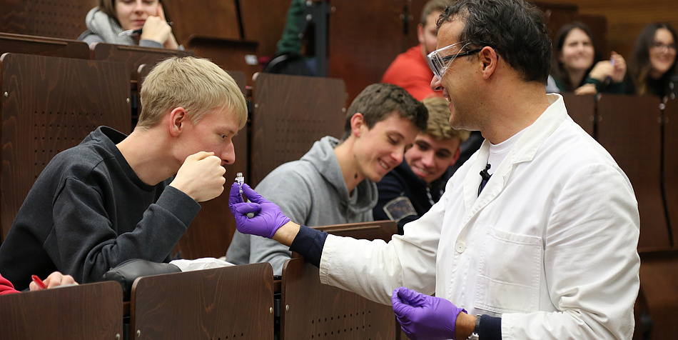 Ein Mann in Labormantel und Handschuhen reicht einem Studenten eine Riechprobe.