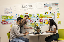 Three people are sitting at a table in front of a wall covered with charts.