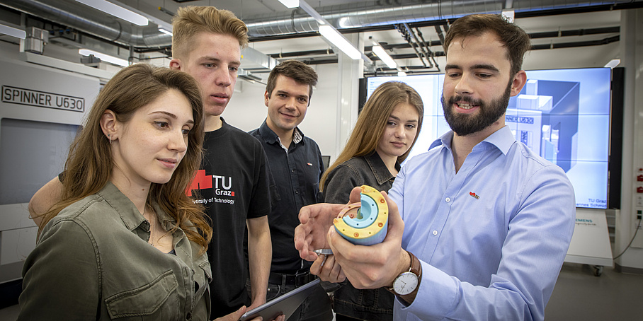 Lehrender erklärt zwei Studentinnen und zwei Studenten ein Maschinenbauteil.