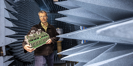 A man stands in a chamber with angular spikes protruding from the wall and holds a large electronic component in his hands.