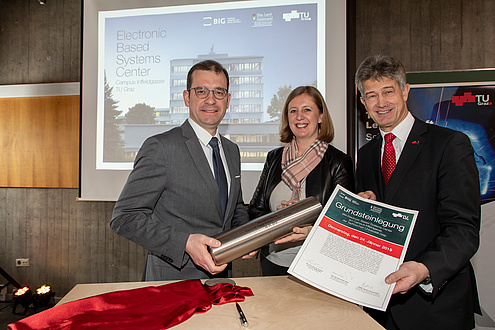 Two men and a woman hold a metal roll and a foundation stone laying certificate in the camera.