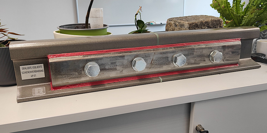 Ein Stück einer Eisenbahnschiene, an deren Seite eine Verstärkung mit Schrauben angebracht ist. In der Mitte ist die Schiene durch ein dünnes Kunststoffteile in zwei Hälften geteilt.