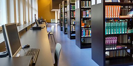 Leerer Gang zwischen Bücherregalen und ebenfalls leeren Arbeitsplätzen in einer Bibliothek