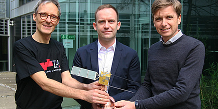 Three men hold a satellite model in their hands and look into the camera.