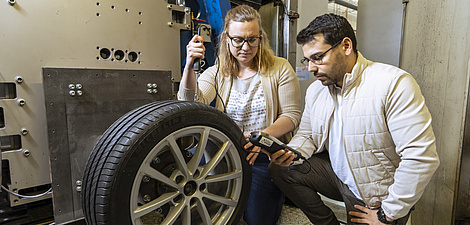 A woman and a man kneel next to a car tyre. The woman puts a measuring device on the tread of the tyre, the man reads the measured result on a display he is holding in his hand.