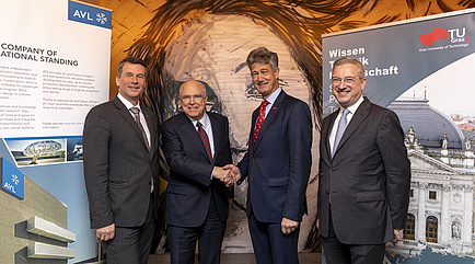 Four men in suits stand in front of the two roll-ups of the AVL and the TU Graz, those in the middle shake their hands. 