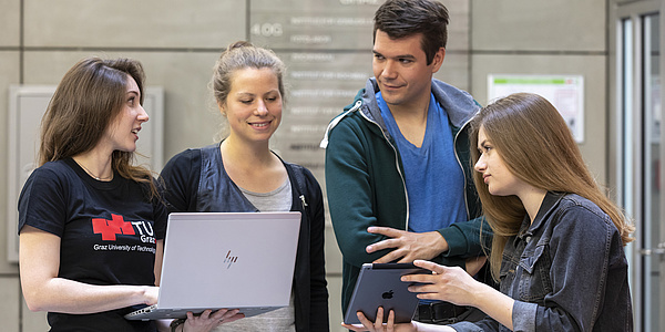 Drei junge Frauen und ein junger Mann blicken auf einen aufgeklappten Laptop und ein Tablet.