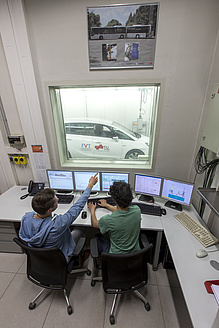 Zwei Personen sitzen vor Bildschirmen und einer Glasscheibe, hinter der ein Fahrzeug zu erkennen ist.