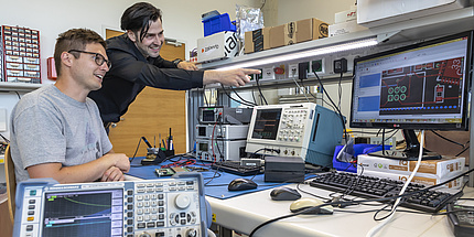 Zwei Männer an einem Tisch mit mehreren elektronischen Geräten, vielen Kabeln und kleinen Bauteilen.