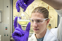 Man in safety glasses and purple rubber gloves holds a glass bulb with bright liquid.