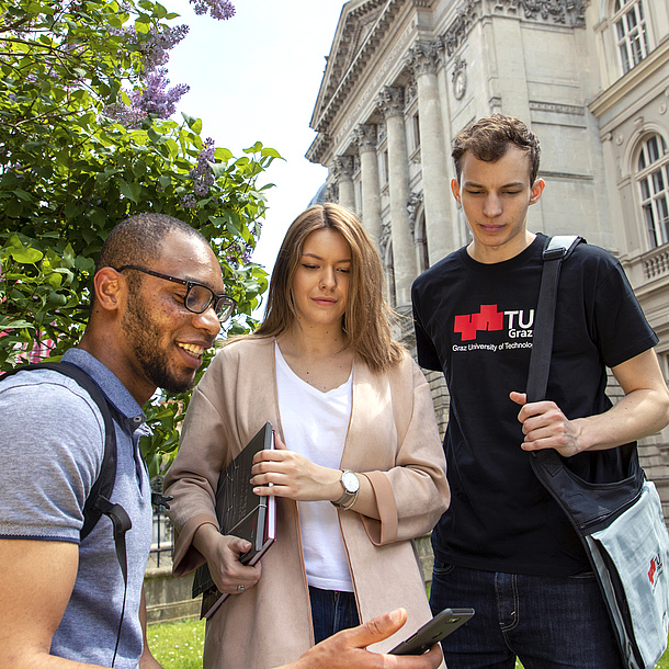 3 Studierende stehen vor der alten Technik und blicken auf ein Smartphone.