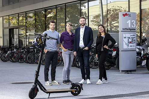 Four people, two women and two men, look into the camera. An e-scooter stands in front of them.