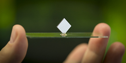 Zwei Finger halten eine Glasplatte auf der ein weißes Quadrat balanciert.