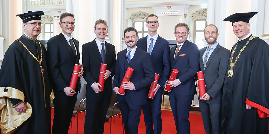 Acht Männer lächeln in die Kamera. Der Mann ganz links und der Mann ganz rechts tragen eine festliche Robe. Die sechs Männer in der Mitte halten rote Dokumentenrollen.