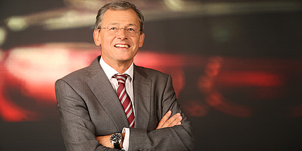 Man in grey jacket with red and silver striped tie, greying hair, fine spectacles, open facial expression and crossed arms in portrait.