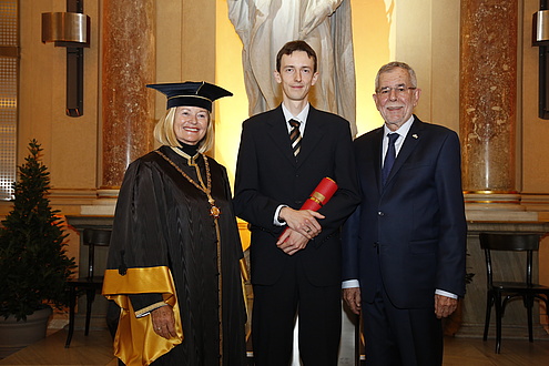 Drei Männer in festlichem Gewand, jener in der Mitte hält eine rote Diplomrolle in der Hand.