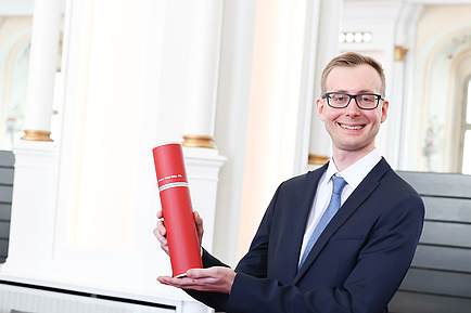 A man holds up a red document roll and smiles into the camera.