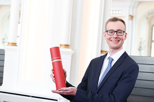 A man holds up a red document roll and smiles into the camera.