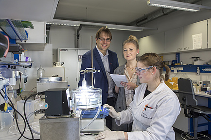 A woman in the lab coat sits in front of a brightly lit apparatus, next to her there is a man and another woman, both of whom look into the camera.