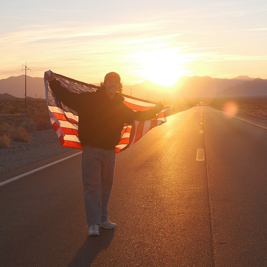 Ein junger Mann mit USA-Flagge steht im Sonnenuntergang inmitten einer Straße.