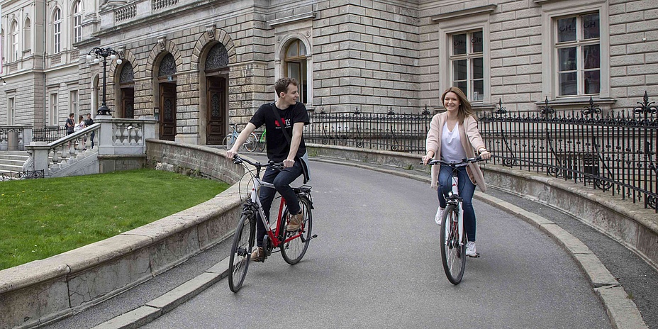Young man and woman on a bicycle on the driveway of a historic building.