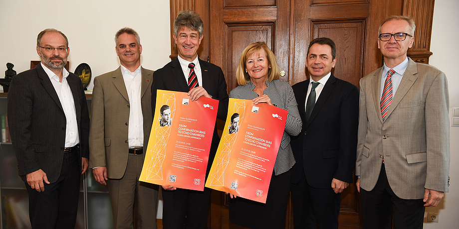 Stefan Vorbach (Professor für Unternehmensführung und Organisation), Dekan Roderick Bloem, Rektor Harald Kainz, Rektorin Christa Neuper, Dekan Thomas Foscht, Studiendekan Heinz Königsmaier stehen vor einer hölzernen Doppelflügeltüre. Rektor Harald Kainz und Rektorin Christa Neuper halten die A3-Werbeplakate zur Auftaktveranstaltung in die Kamera.