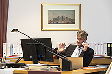 Mann mit dunklen Haaren sitzt am Schreibtisch, telefoniert und winkt. 