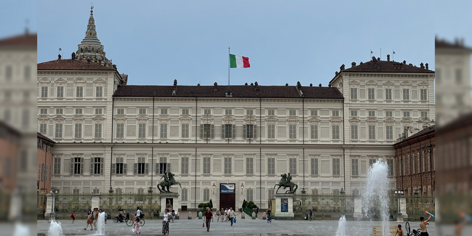View of the Palazzo Reale, a baroque palace made of beige stone in front of a square surrounded by water columns.