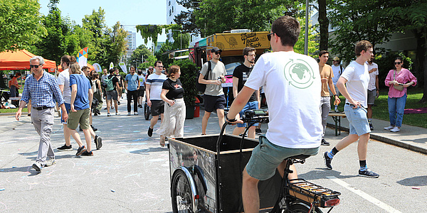 Eine Person in weißem T-Shirt fährt auf einem Lastenrad eine Straße entlang. Am Straßenrand stehen grüne Bäume, ein Foodtruck und einige Fahnen. Mehrere Personen gehen die Straße entlang.