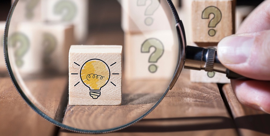 Holzwürfel mit Fragezeichen, Hand mit Lupe findet Holzwürfel mit Glühbirnen-Symbol