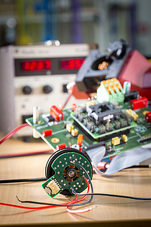 Ein Elektromotor liegt auf einer Tischplatte, im Hintergrund eine Platine.