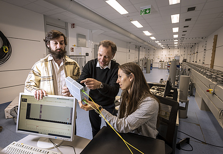 Three people look at a box, the person in the middle is pointing at it.