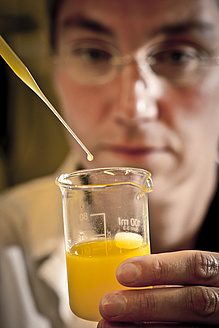 Close-up of a pipette taking orange liquid from a glass beaker, behind which a woman can be seen.