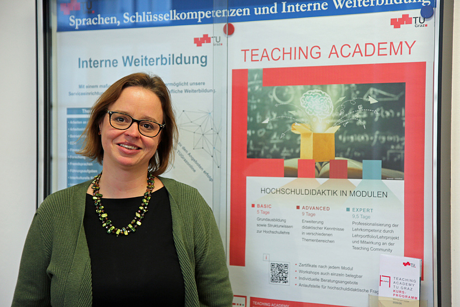 Eine Frau mit kinnlangen Haaren steht neben einem Plakat der Teaching Academy. Sie trägt ein schwarzes Shirt und einen grünen Cardigan.